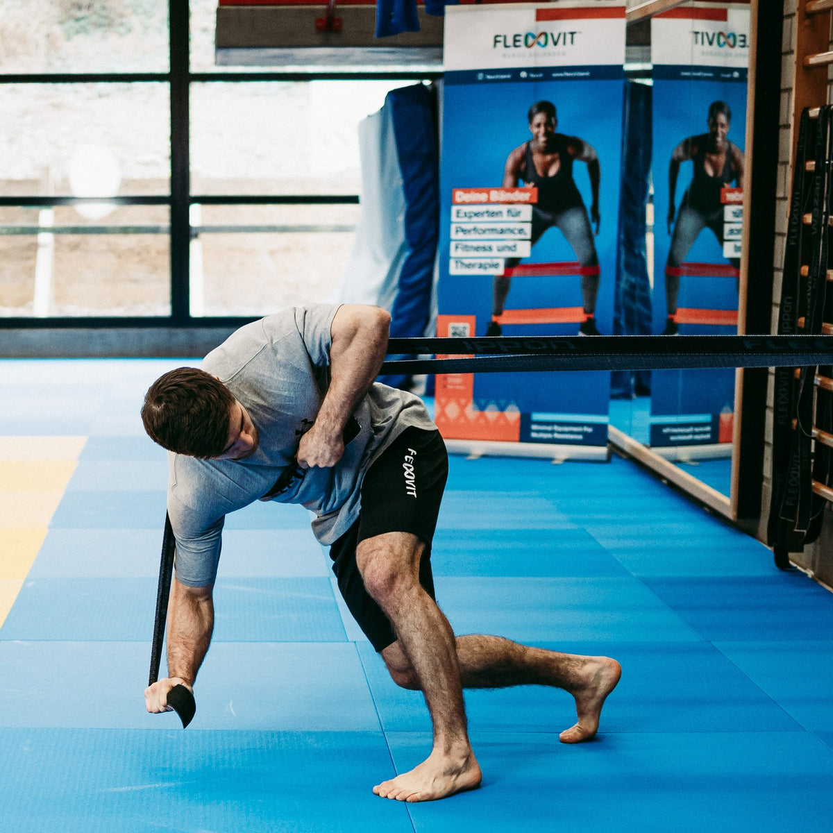 Hombre descalzo sobre una colchoneta azul tira de una banda negra, Flexvit Sparring